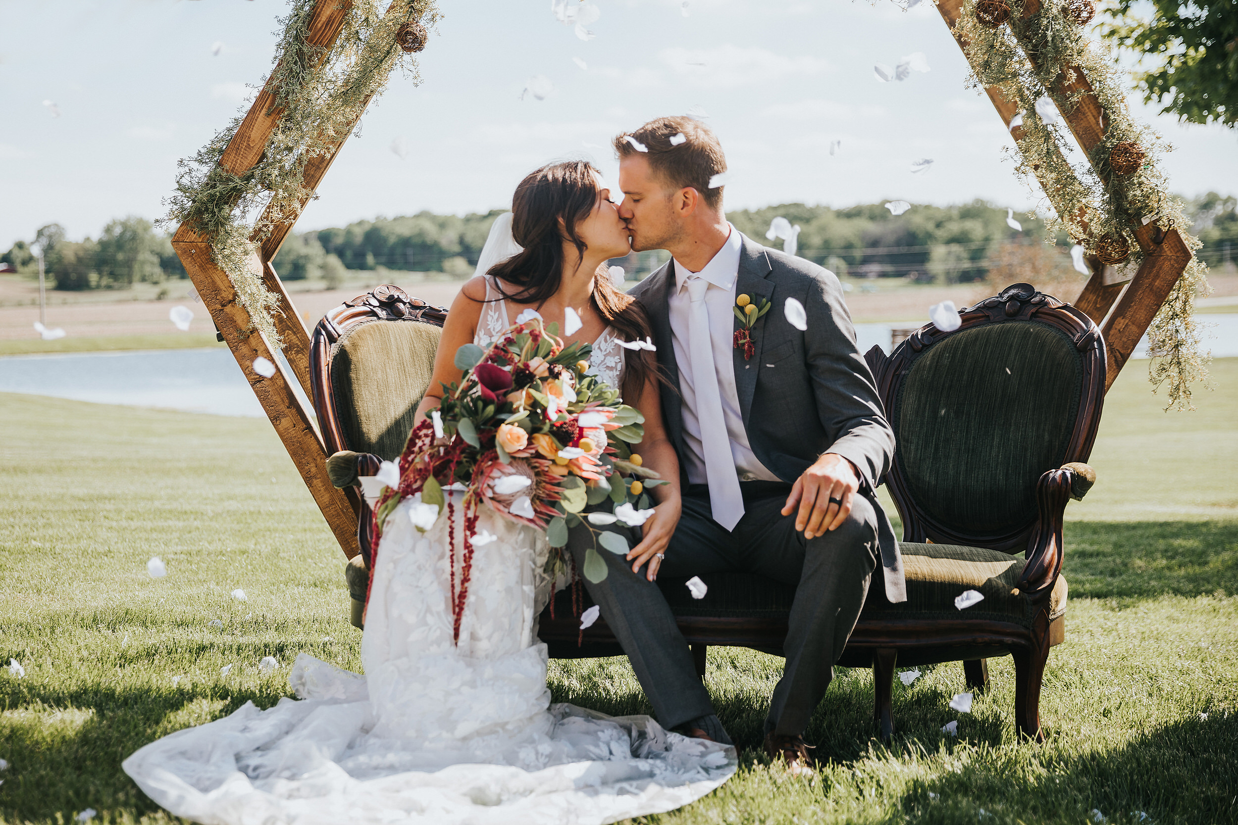 A bride and groom kiss while sitting on a green victorian styled couch under their wedding arbor. Flower petals fall around them.
